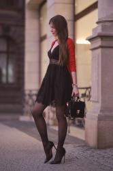 Czarna koronkowa sukienka, czerwony cardigan, czarne rajstopy i lakierowane szpilki